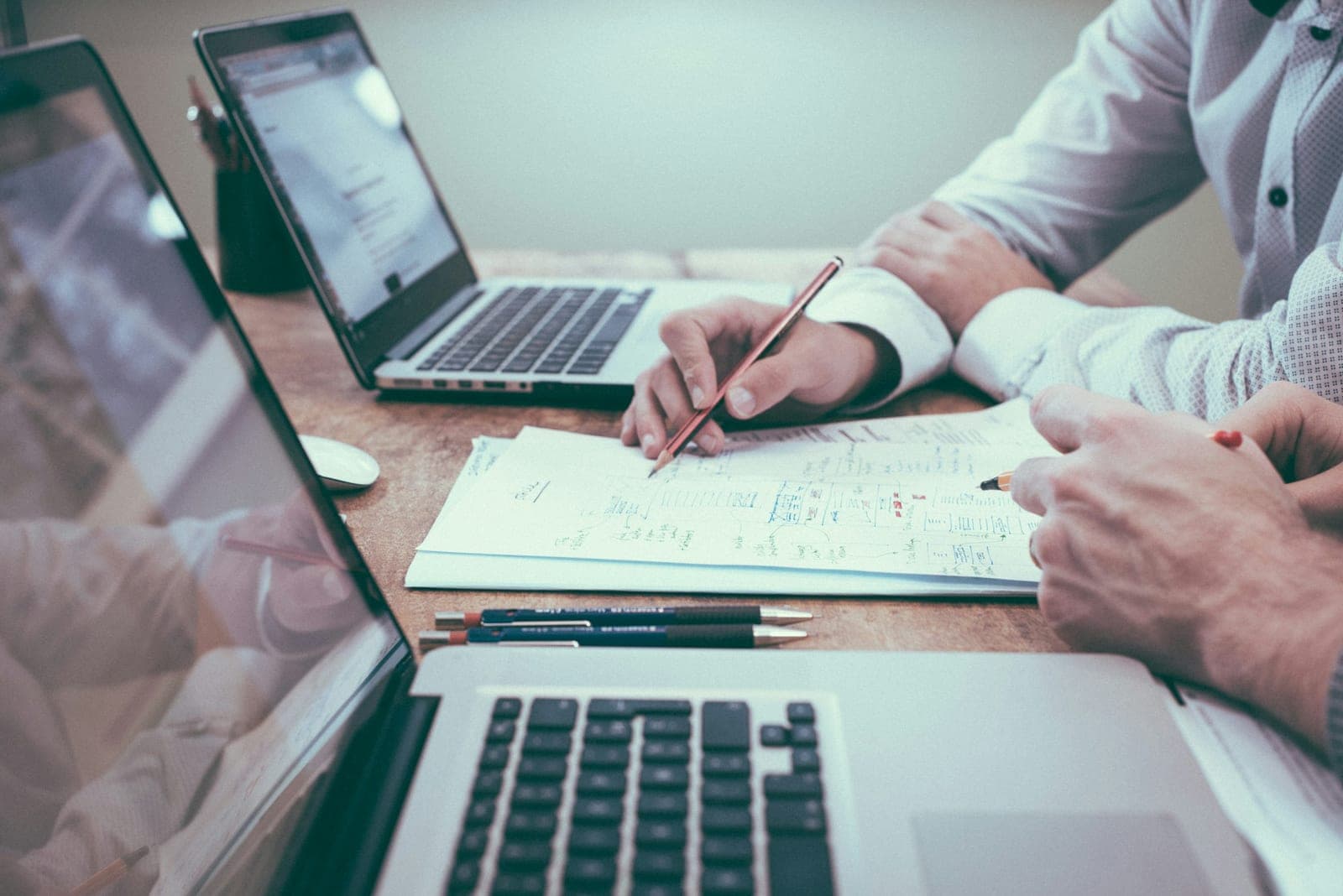 Hands of two people reviewing annotated documents with pencils beside two open laptops on a wooden table