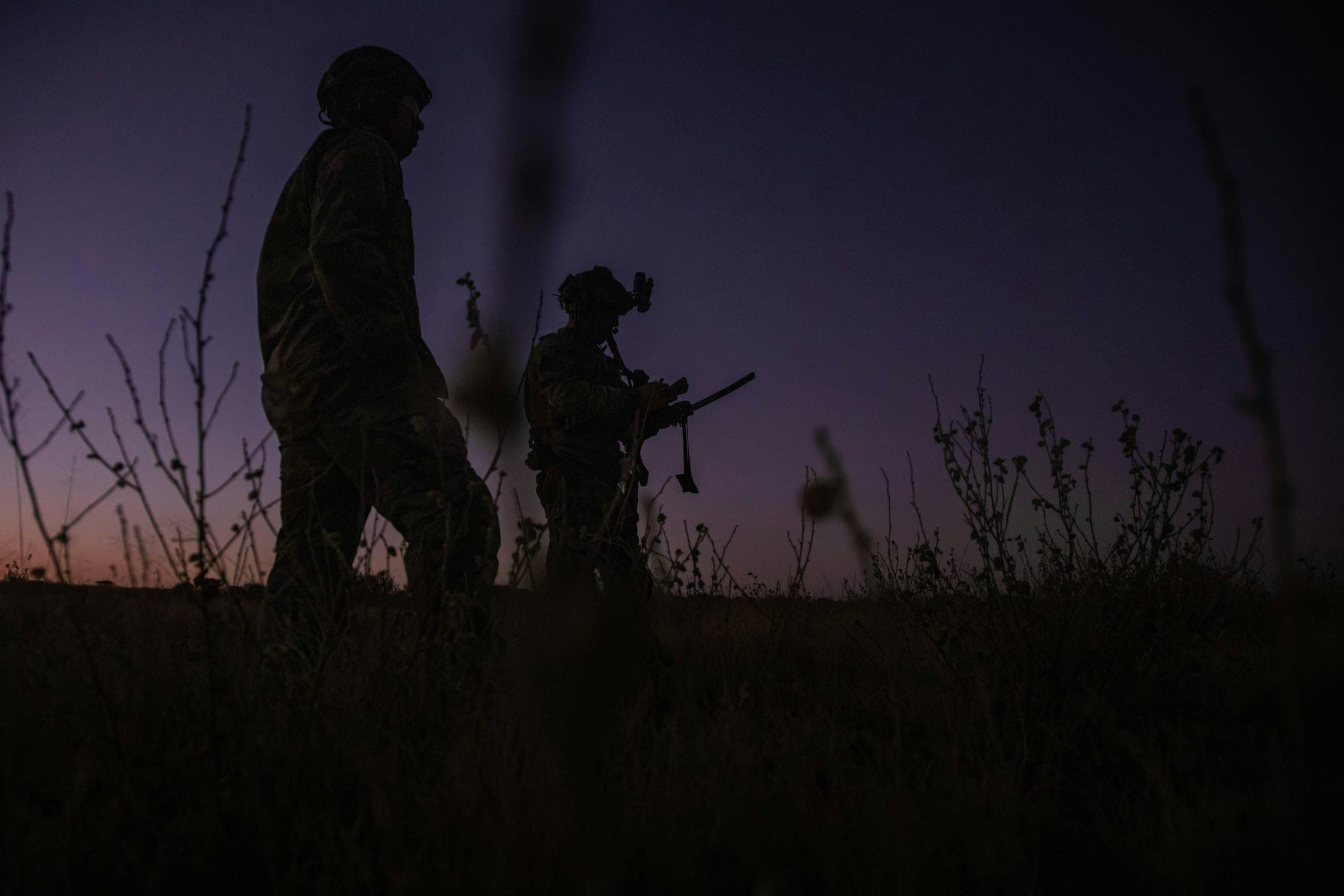 Two helmeted soldiers in silhouette walking through tall grass at dusk, one with a night-vision mount