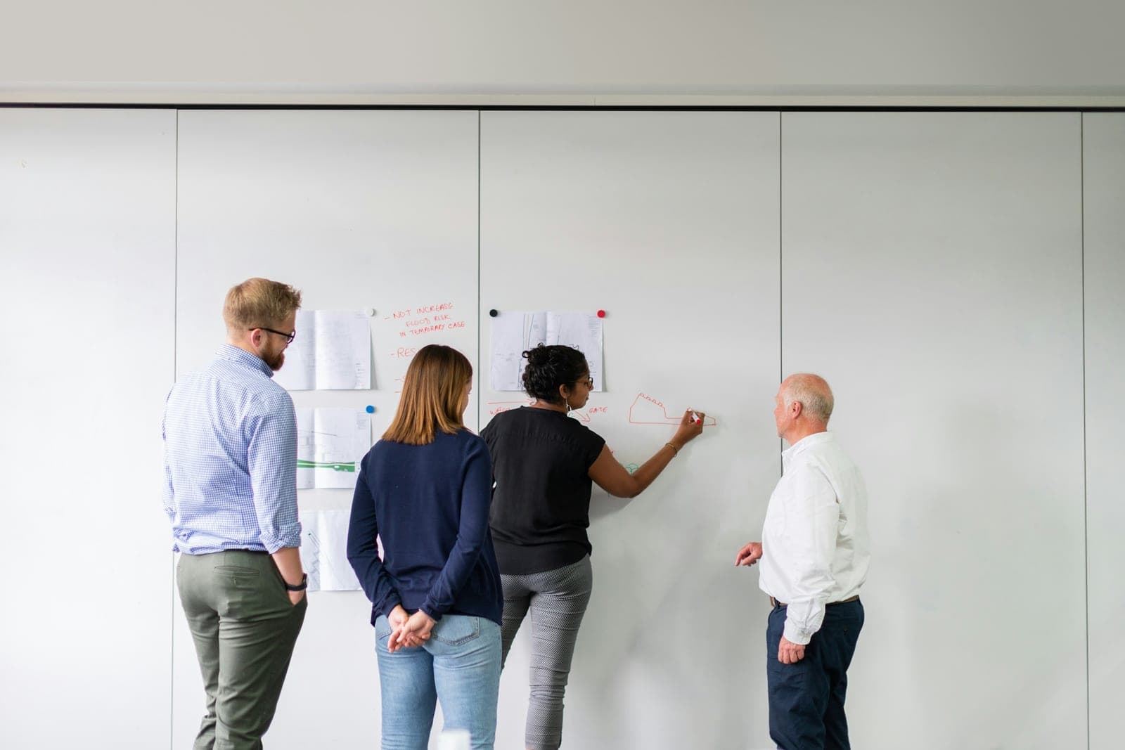 Four colleagues seen from behind and the side at a whiteboard with pinned technical drawings as one sketches a plan
