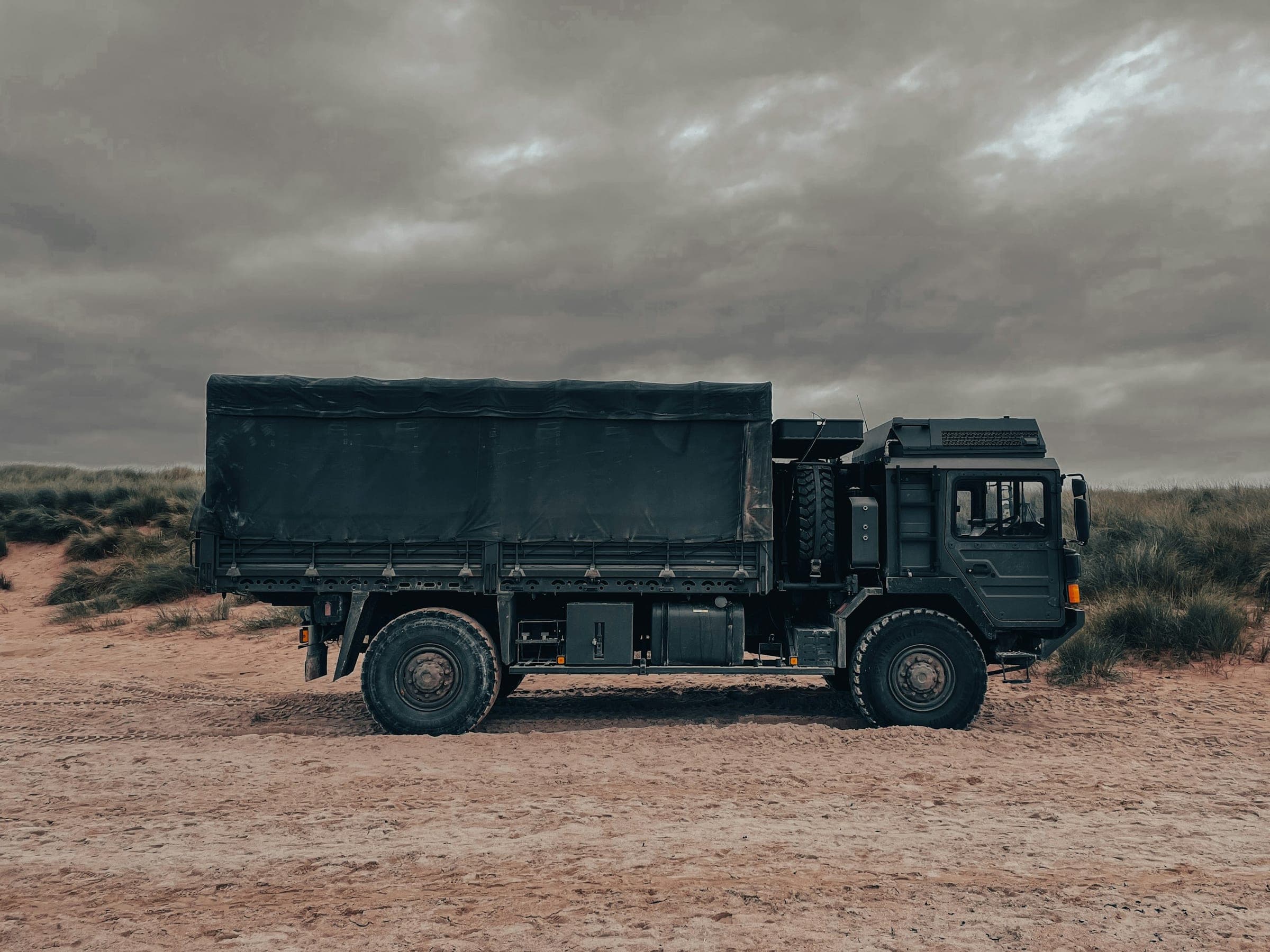 Dark green military cargo truck with a canvas cover parked on sandy ground under a cloudy sky