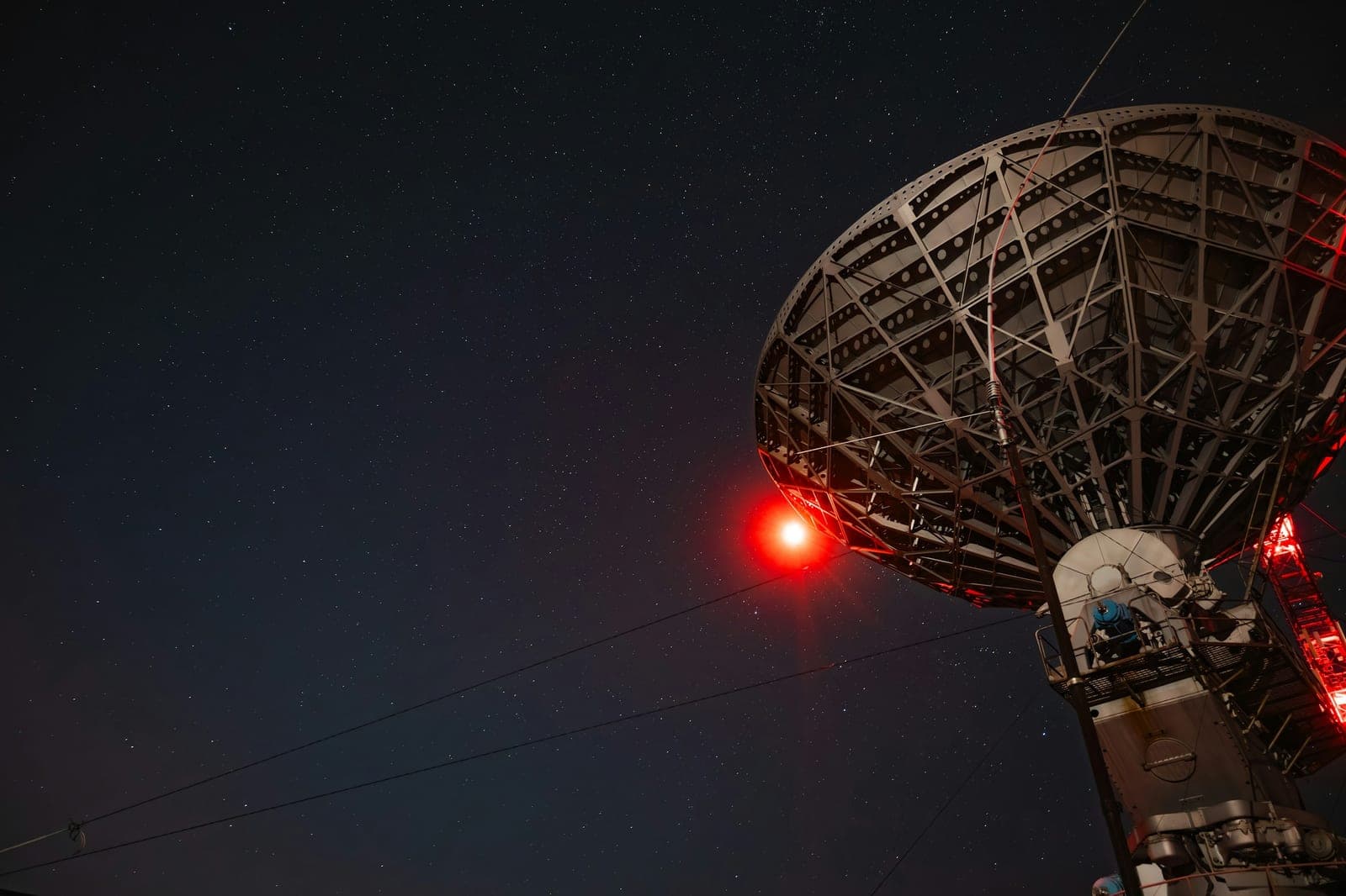 Large dish antenna and its mount lit by red warning lights against a starry night sky, seen from below