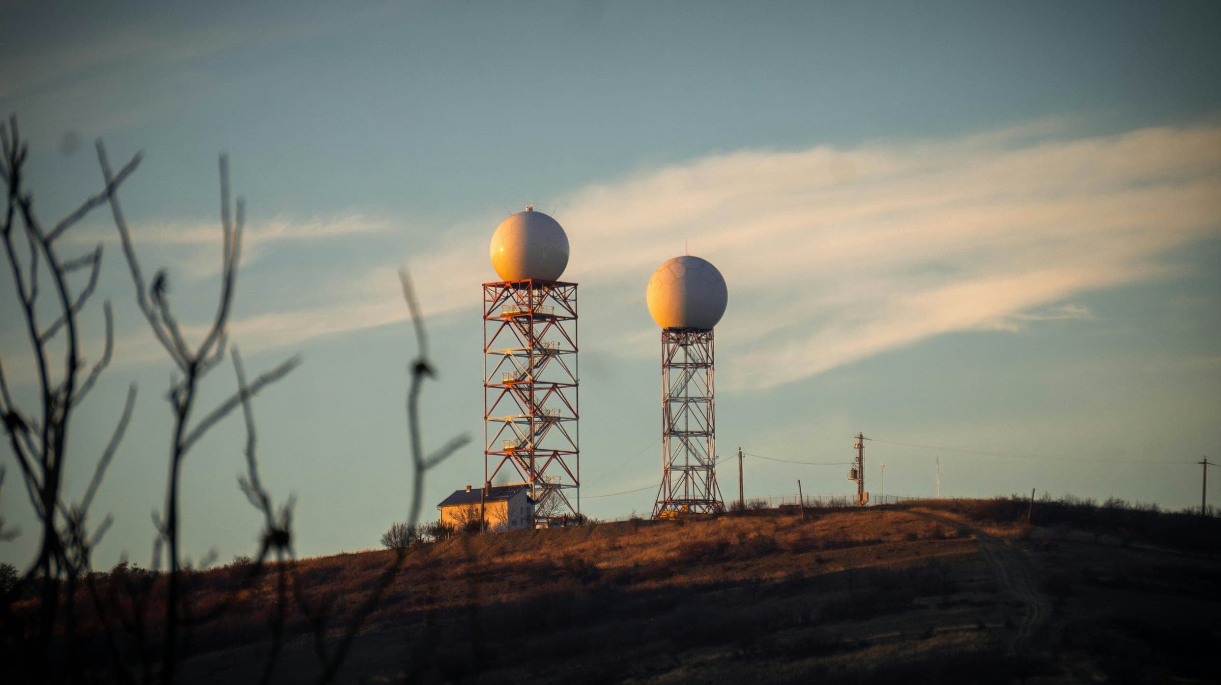 Two radar domes on lattice towers at a fenced hilltop station, lit by low golden evening sun