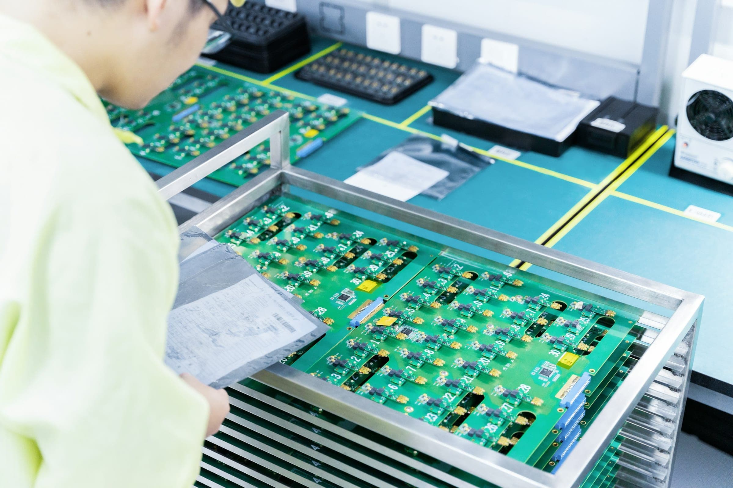 Worker's arm beside trays of green circuit boards on an electronics production line