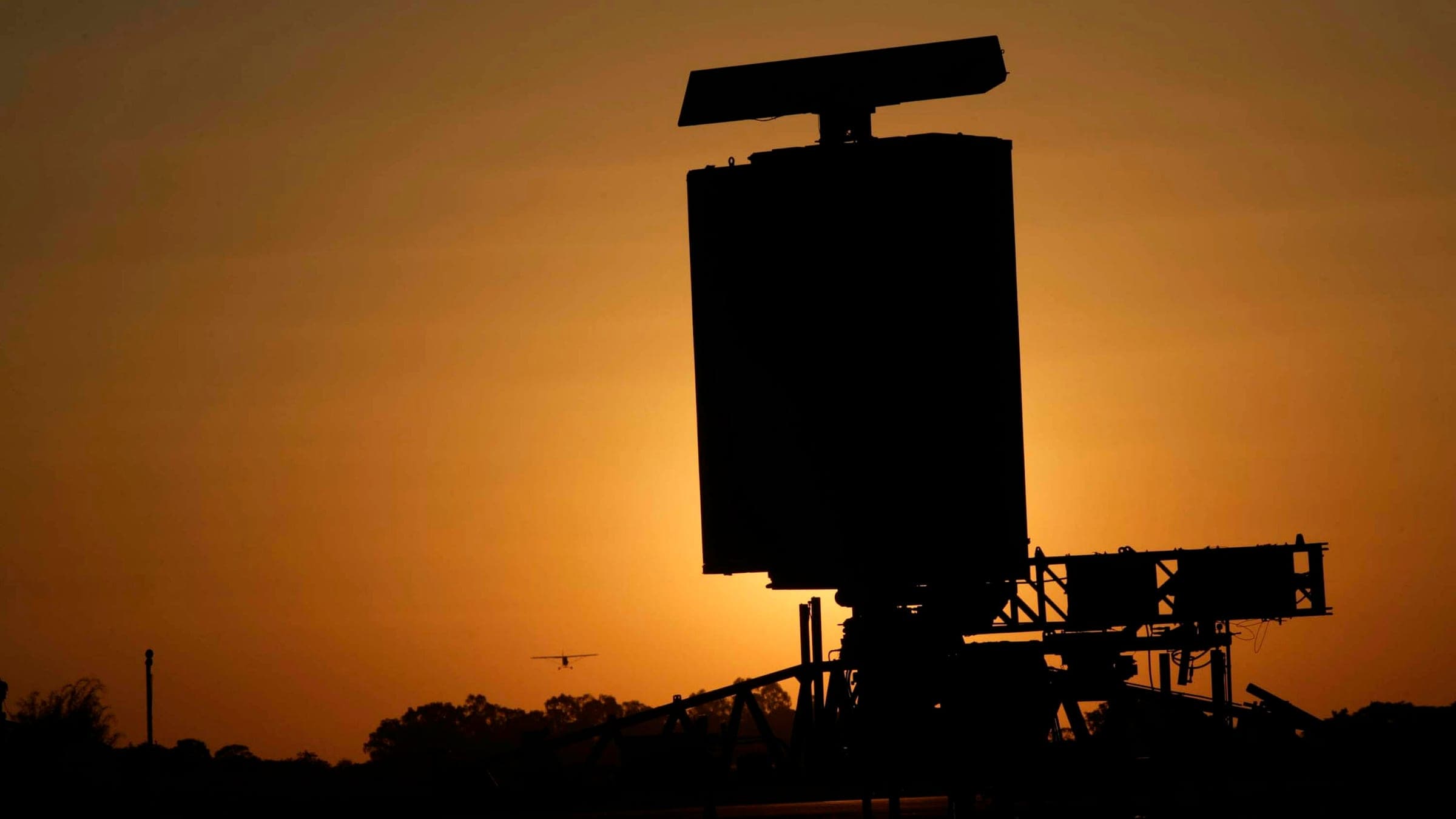 Silhouette of a surveillance radar antenna on its mount against an amber sunset sky, small aircraft in the distance