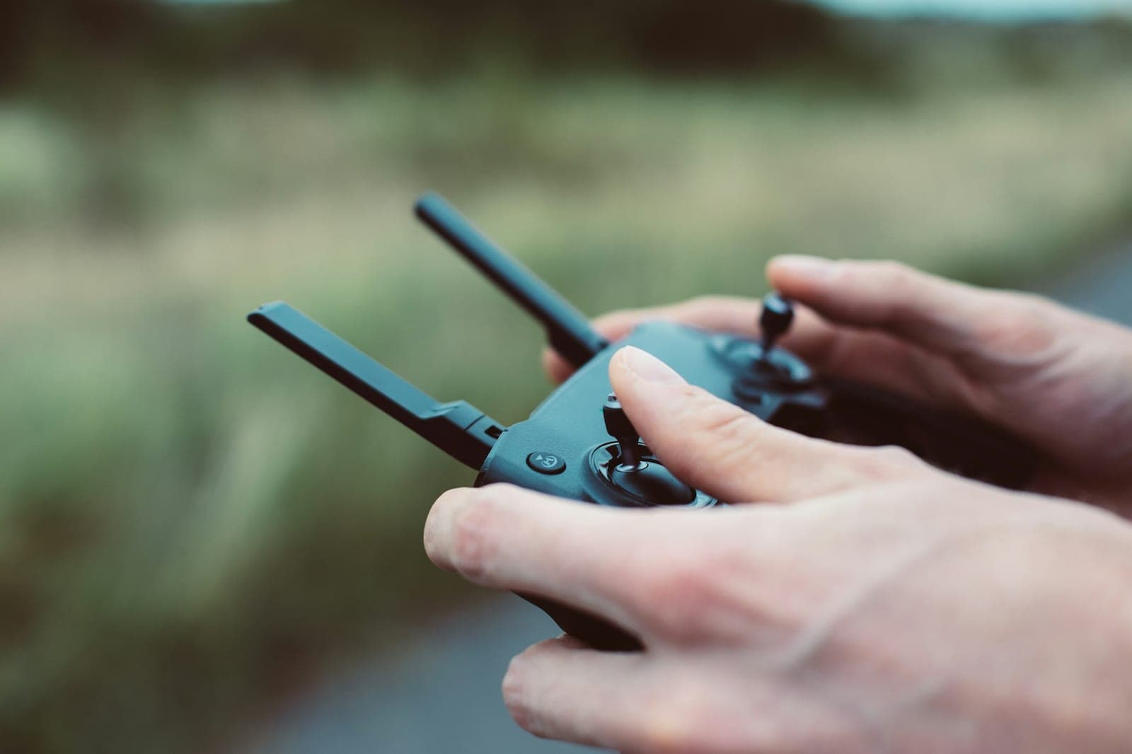 Two hands working the control sticks of a drone remote controller outdoors, muted green field blurred behind