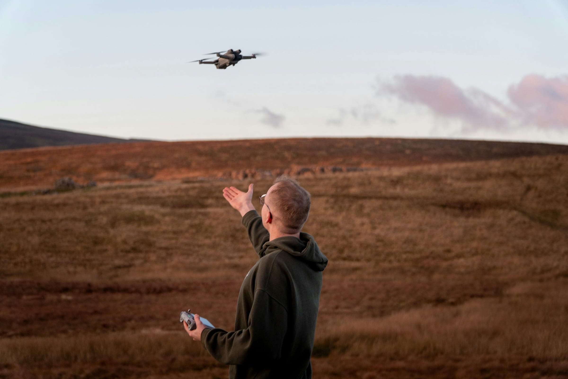 Operator seen from behind sends a small drone off by hand over russet moorland at dusk, controller in his other hand